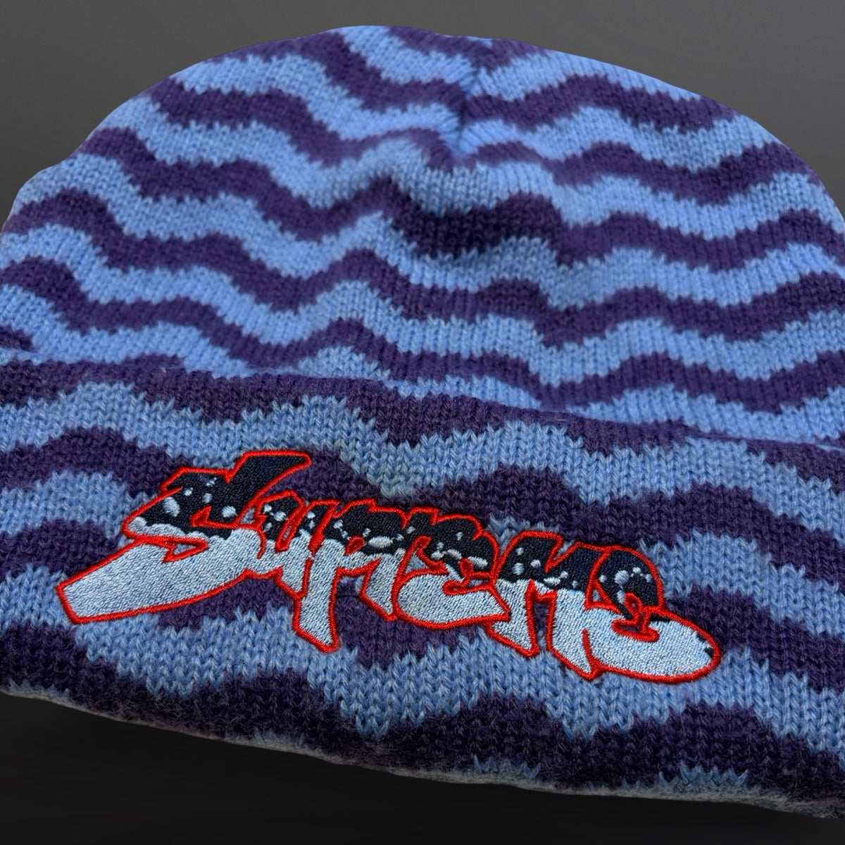 Detalje af Supreme-logo i sort og lyseblåt med rød kontur på blå og lilla mønstret hue